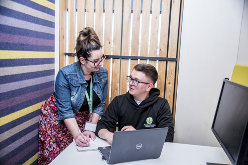 Two members of staff are looking at each other and are in conversation. Becca is writing on a notepad and Kieran has a laptop in front of him.