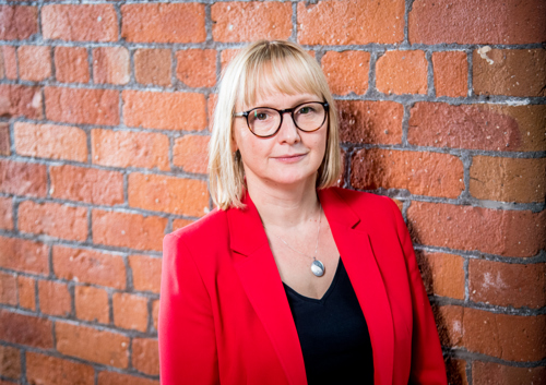 Jules, our Director of Resources is stood against a brick wall smiling at the camera. She has a blonde bob, wearing glasses and wearing a red blazer.